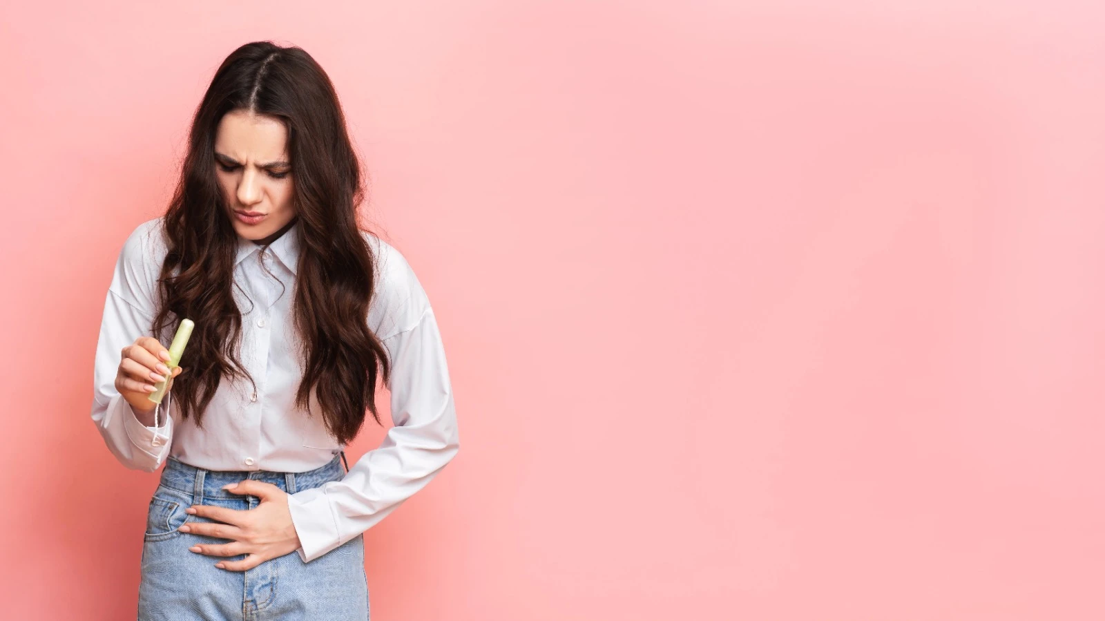 Young woman holding a tampon and touching her stomach, experiencing menstrual cramps and PMS/PMDD symptoms on a pink background